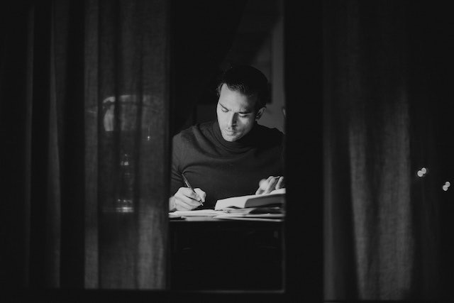 black and white - man writing at a desk behind some half-open curtains.